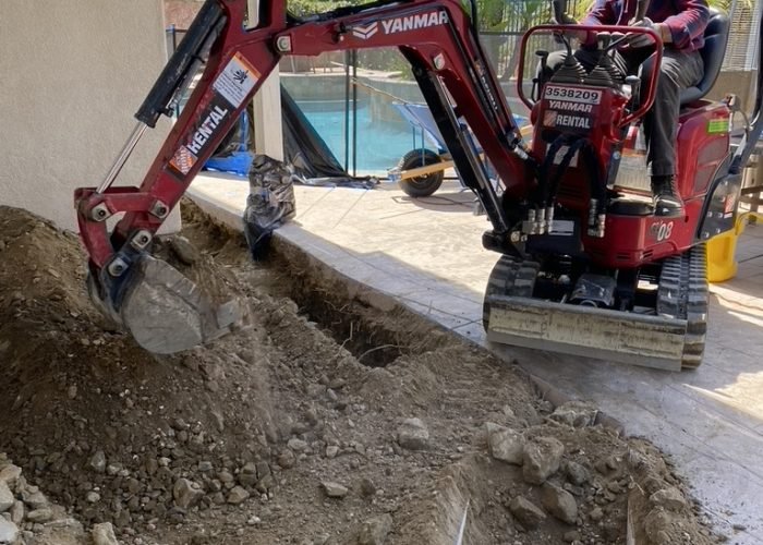 Mini-excavator trenching for a new sub-grade plumbing line near a residential pool and house foundation.